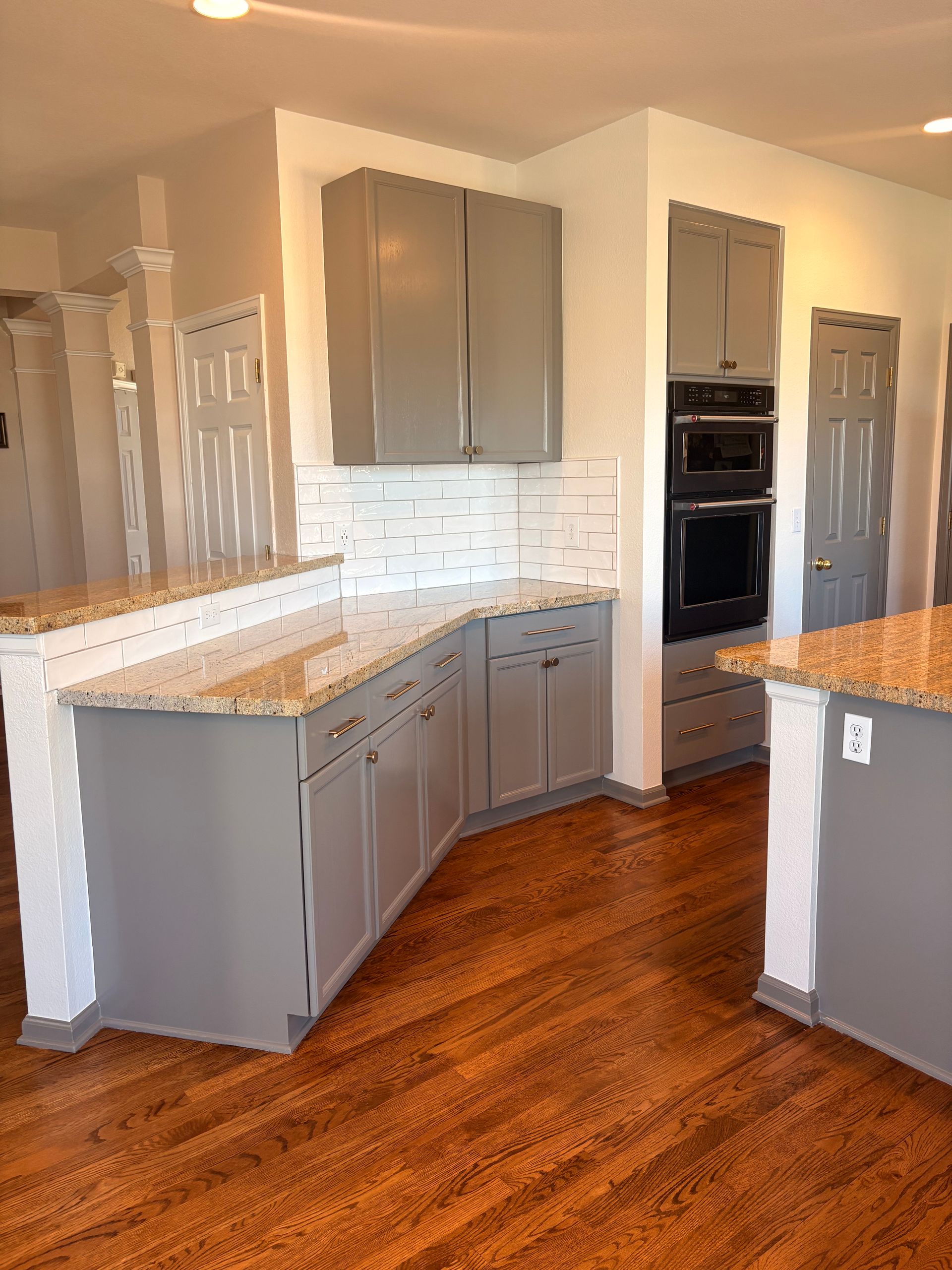 A kitchen with grey cabinets, speckled granite countertops, white tile backsplash, and dark hardwood floors.