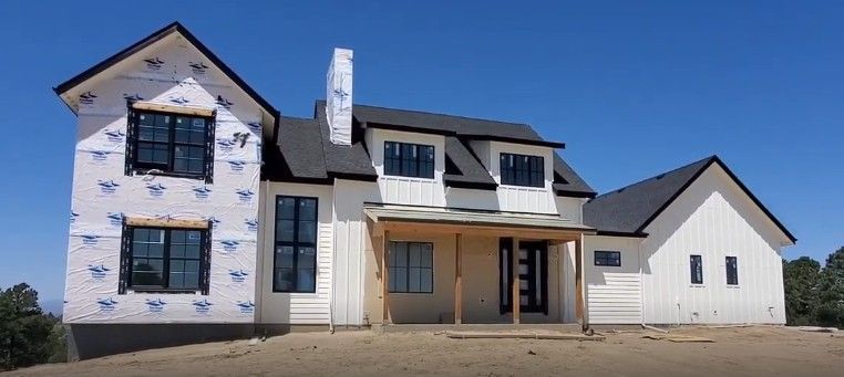 Modern two-story house under construction with black windows and trim, white siding, and a covered porch against a blue sky.