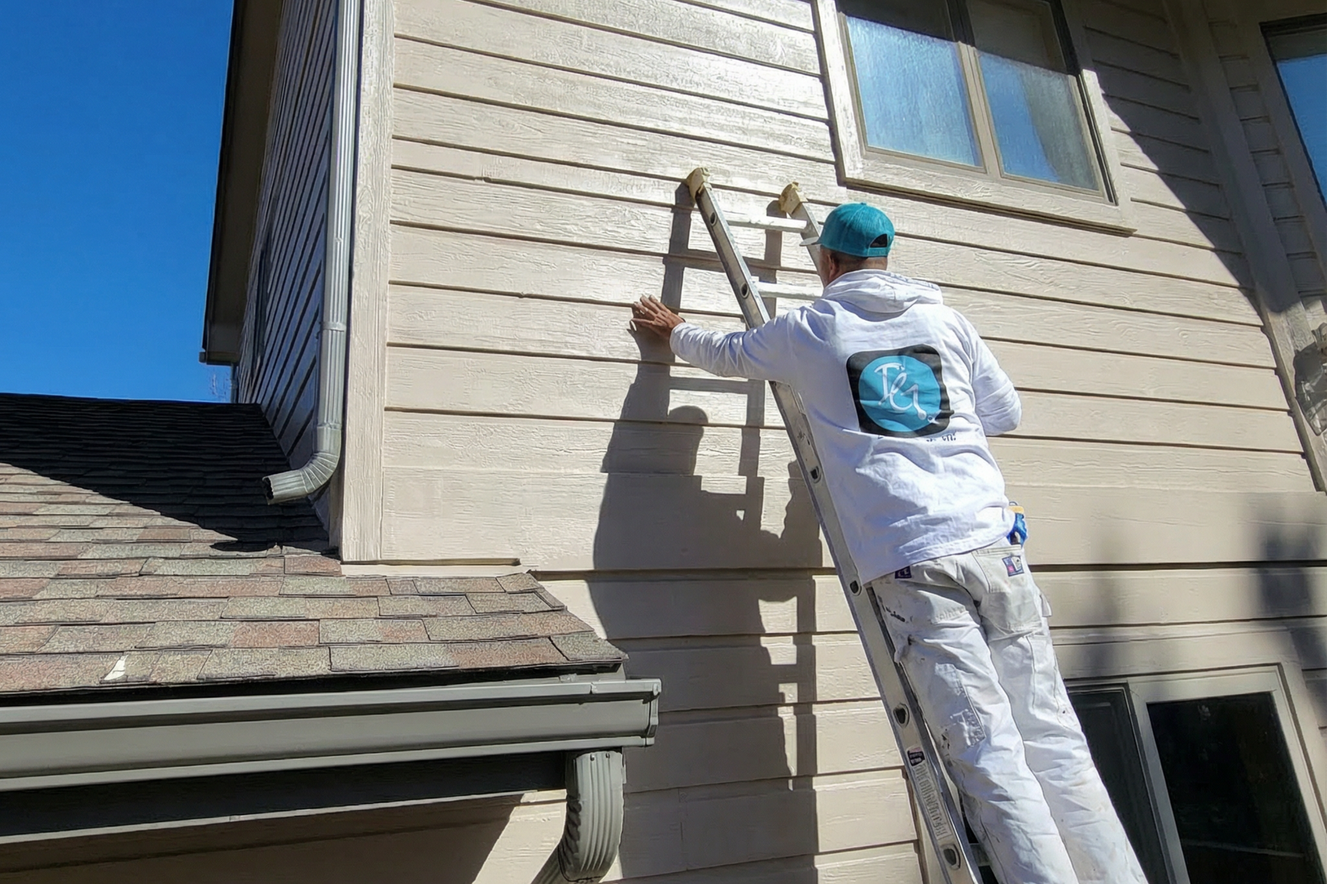 Person on ladder painting house siding, blue sky, tan siding, brown roof, sunny day.