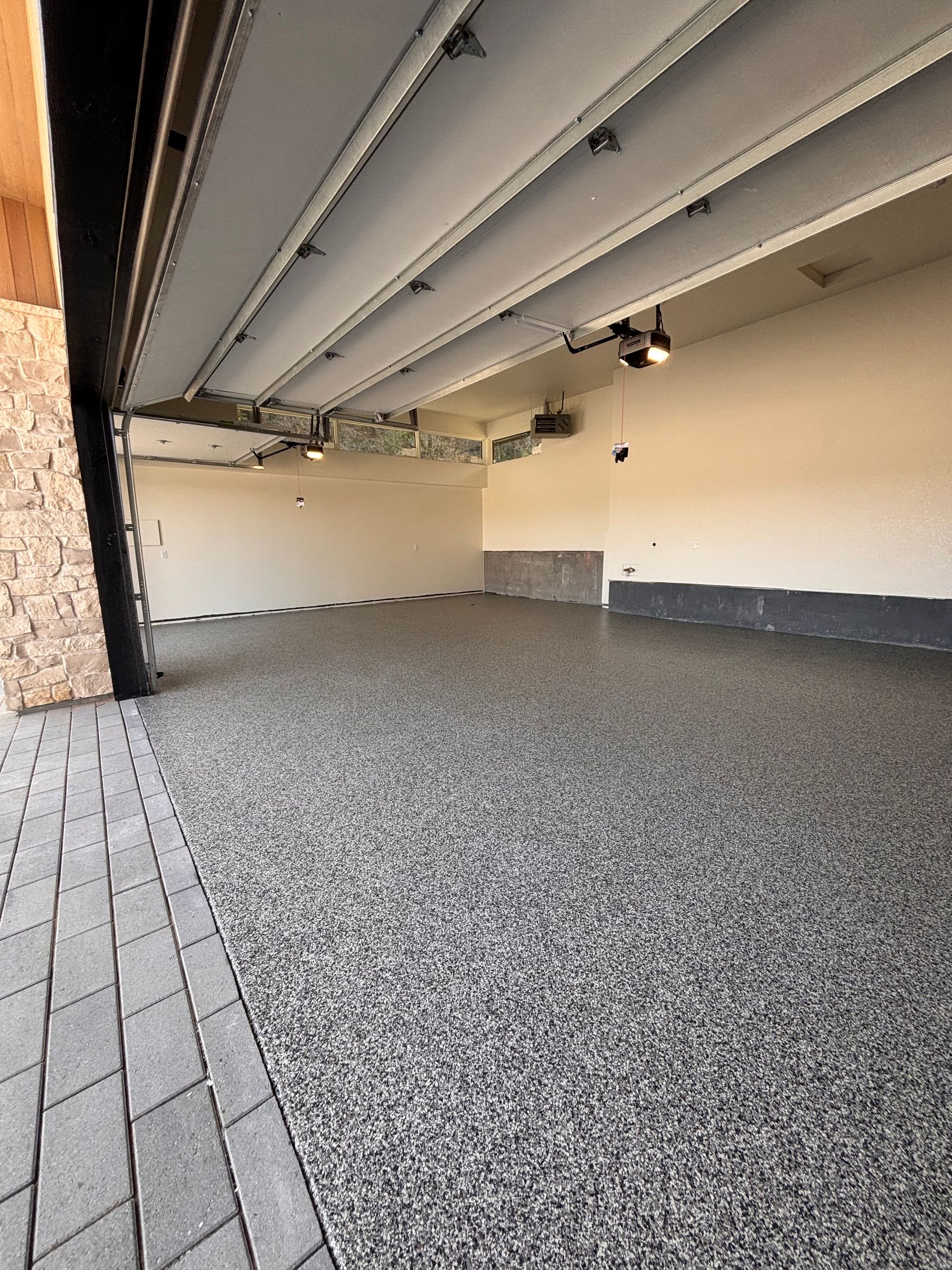 Empty garage with speckled epoxy floor, open door, light-colored walls and ceiling, and stone-paved driveway.
