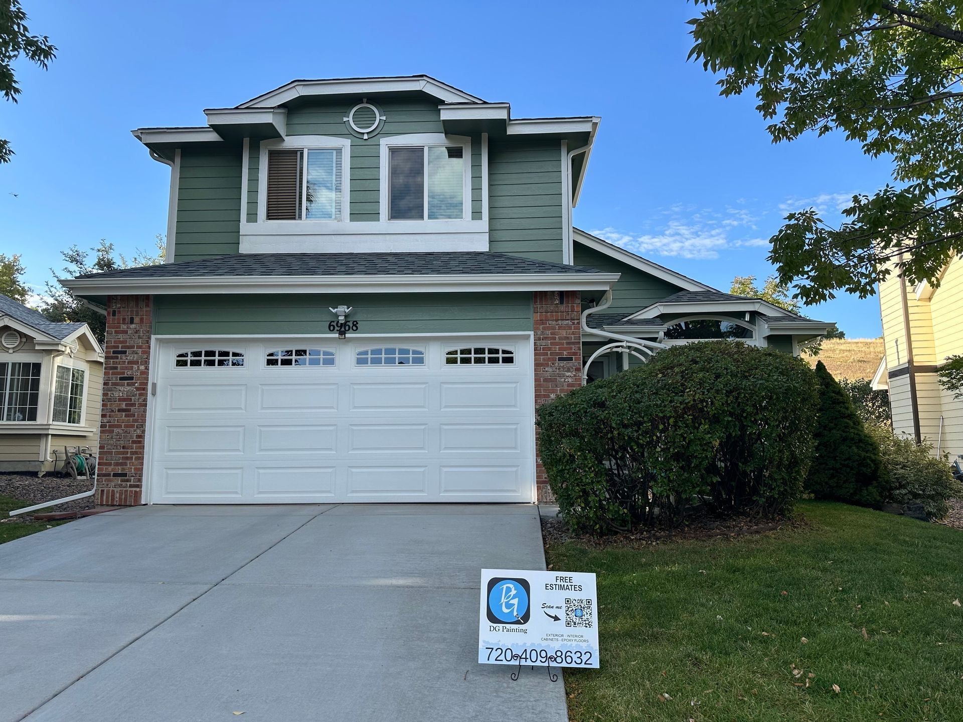 A green house with a white garage door and a sign in front of it.