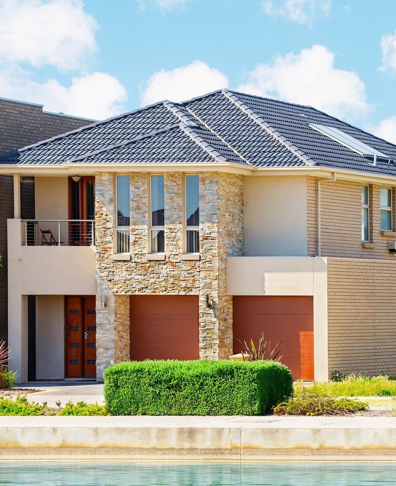 Two-Story Modern Home With Stone and Tan Brick Facade, Brown Garage Doors, and a Green Hedge in Front of a Body of Water — Campbell Built in Newcastle, NSW