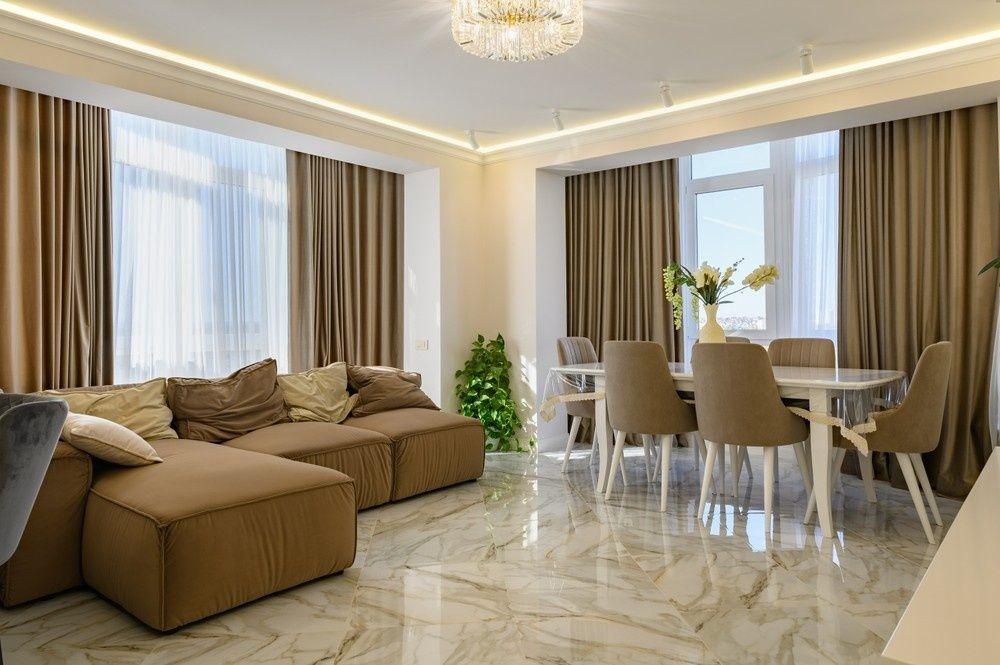 A Modern Living Room With a Brown Sectional Sofa, Dining Table, and Large Windows With Beige Curtains. the Floor is Marble — Campbell Built in Newcastle, NSW