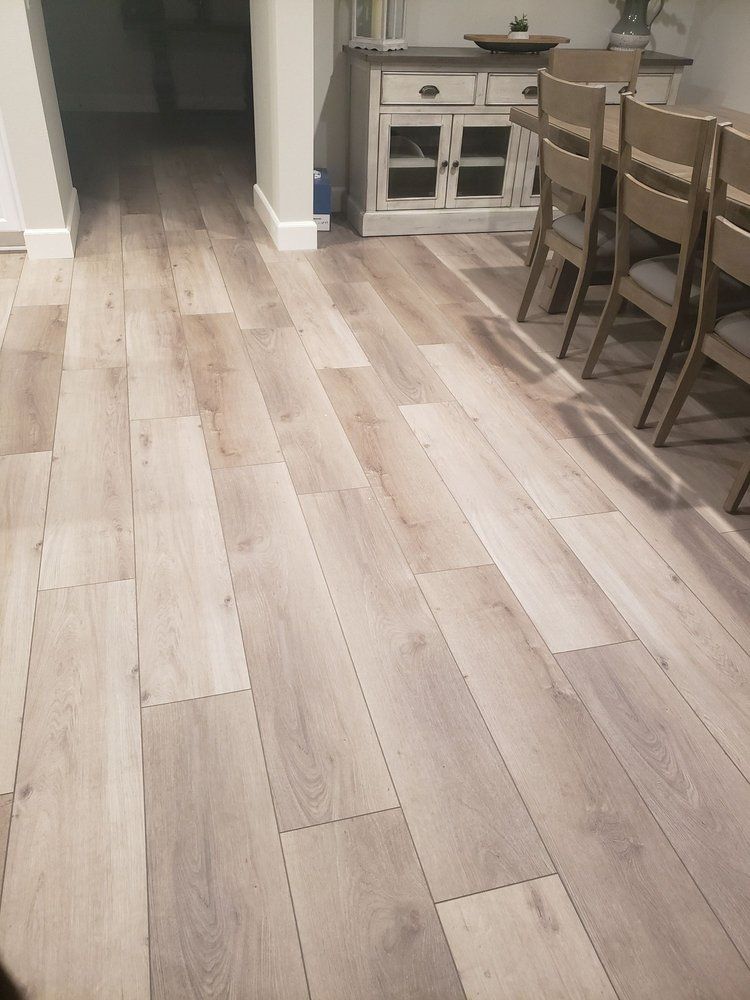 Light-toned wood-look laminate flooring in a home interior with a dining set and wooden cabinet in the background.