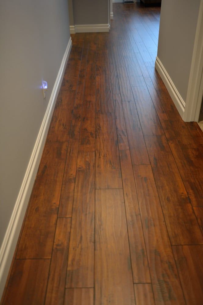 A narrow hallway with warm-toned, brown wood-look plank flooring, light gray walls, and white baseboards.