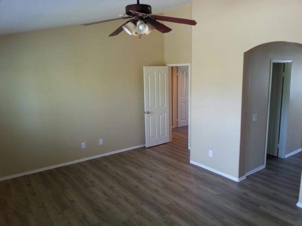 Empty room with beige walls, a ceiling fan, wood-look flooring, an open doorway, and a hallway leading to another room.