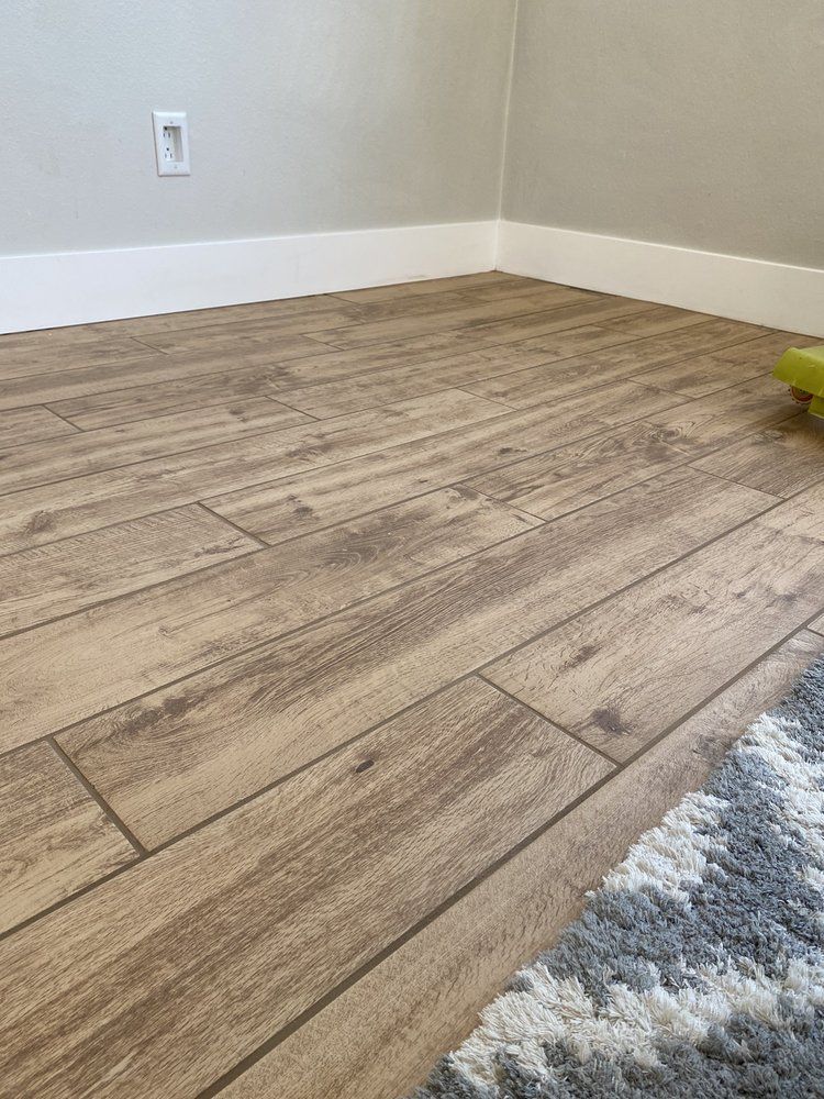 Light brown wood-look plank flooring meets white baseboards in a room corner, with a gray and white rug in the foreground.