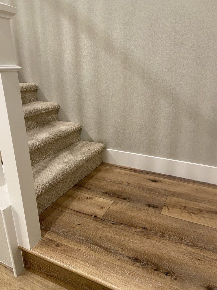 A beige carpeted staircase transitions to wood-look flooring with white baseboards against a light gray wall.