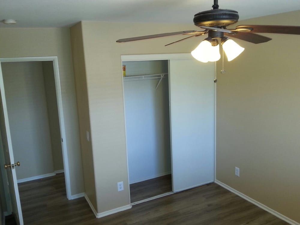 An empty bedroom with beige walls, wood flooring, a ceiling fan, an open closet, and a doorway leading to another room.