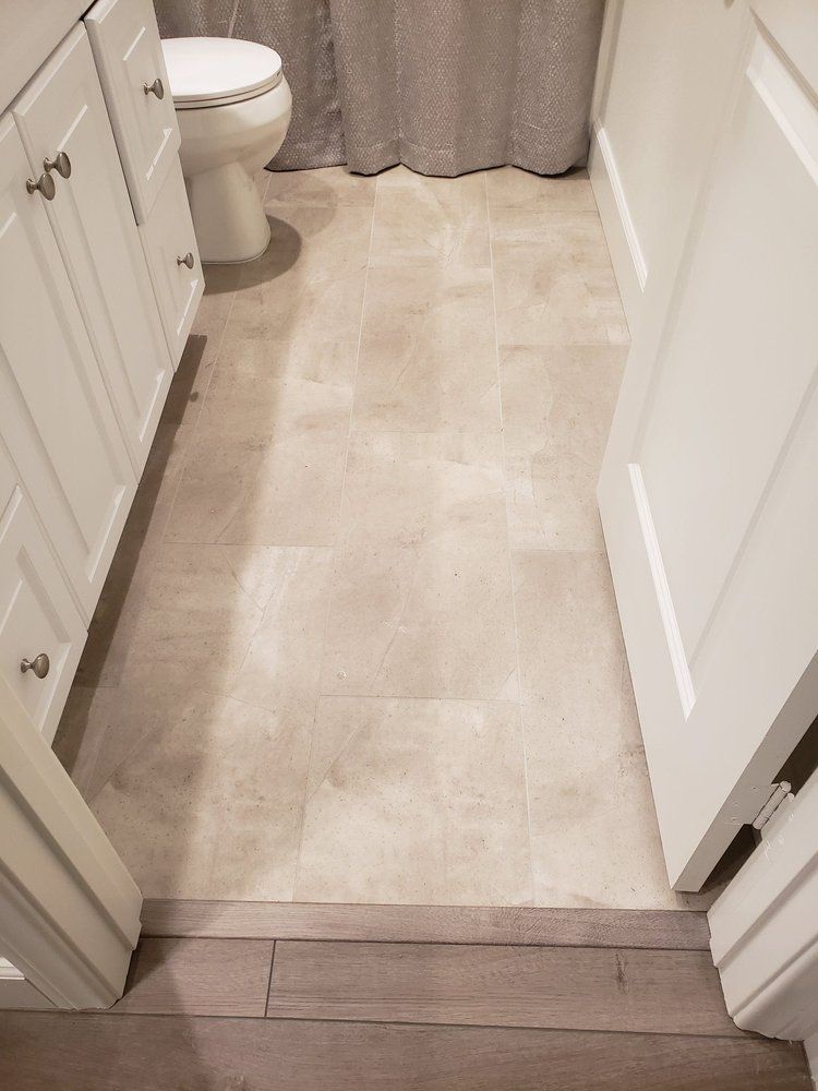 A bathroom with light-colored tile flooring, a white vanity cabinet, a toilet, and a grey curtain, bordered by wood flooring.