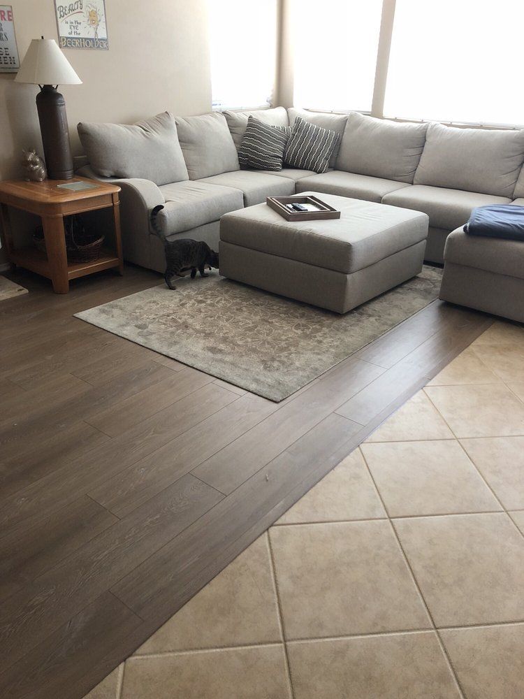 A modern living room with a gray sectional sofa, ottoman, and area rug, featuring a wood-plank floor next to tile.