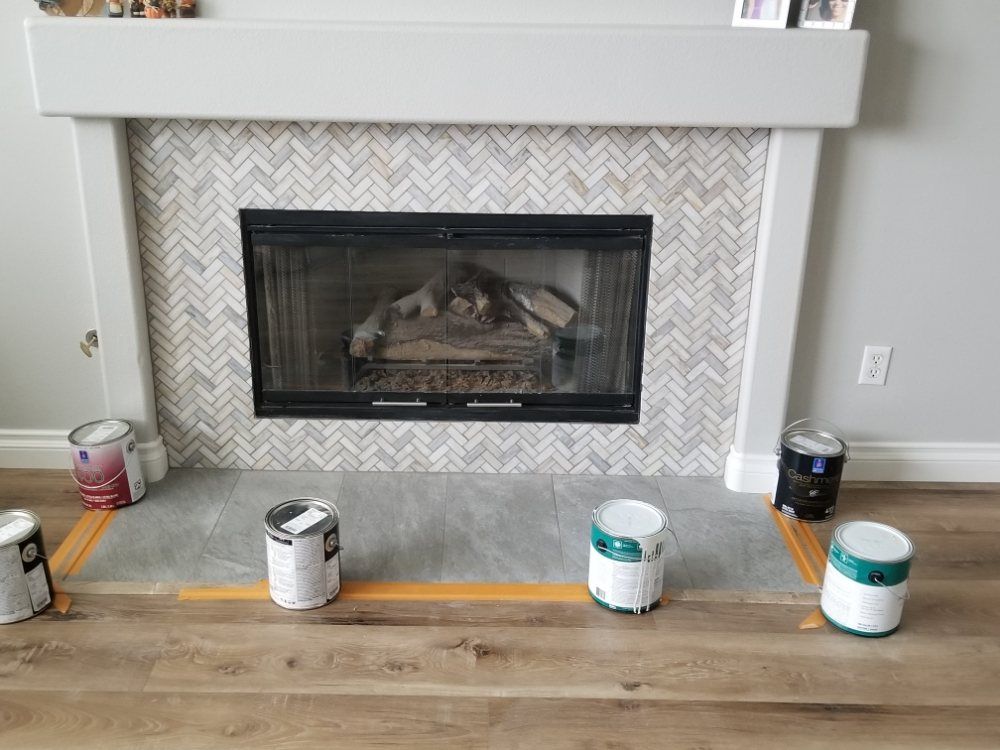 A fireplace with a white mantel and herringbone tile surround, with four paint cans placed on the floor for a project.