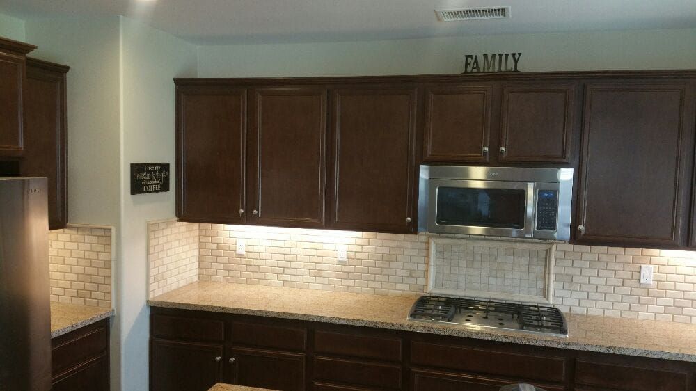 A kitchen featuring dark wood cabinets, a speckled beige countertop, a light-colored tiled backsplash, and a microwave.