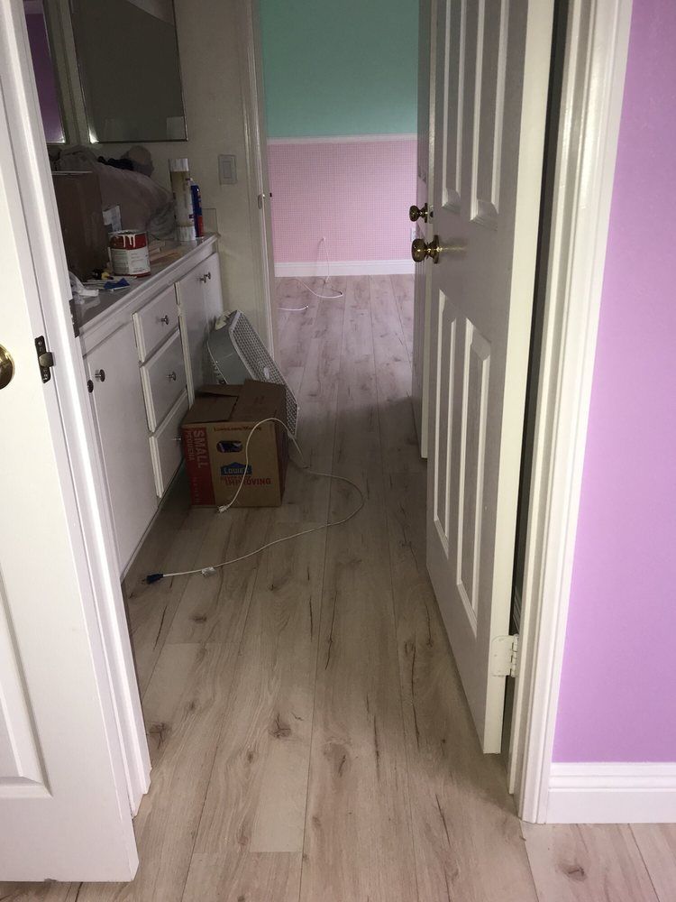 A hallway with light wood-look flooring connects a bathroom with white cabinets to a room with pink and green walls.