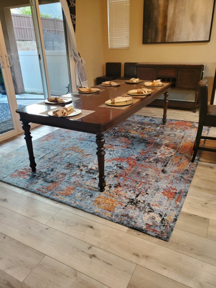 A dark wooden dining table set for six on a large, multi-colored patterned rug in a bright, modern dining room.