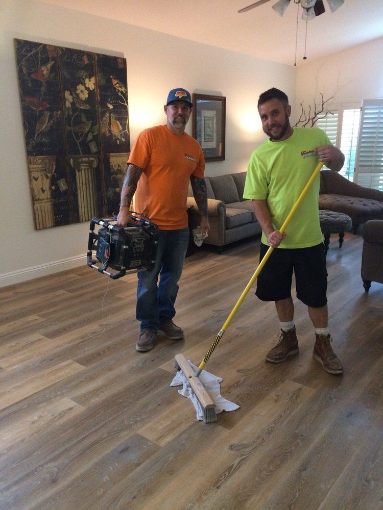 Two workers in bright shirts clean a living room, one holding a floor tool and the other carrying equipment.