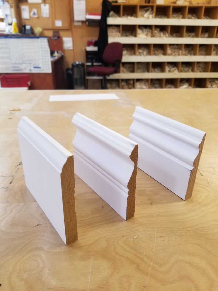 Three samples of white, pre-primed baseboard molding stand upright on a wooden workshop table.
