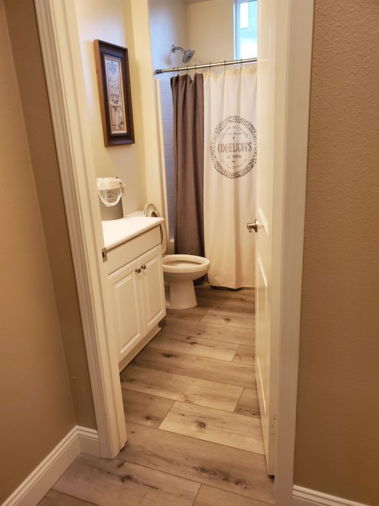 A doorway view of a small bathroom with wood-look flooring, a white vanity cabinet, a toilet, and a beige shower curtain.