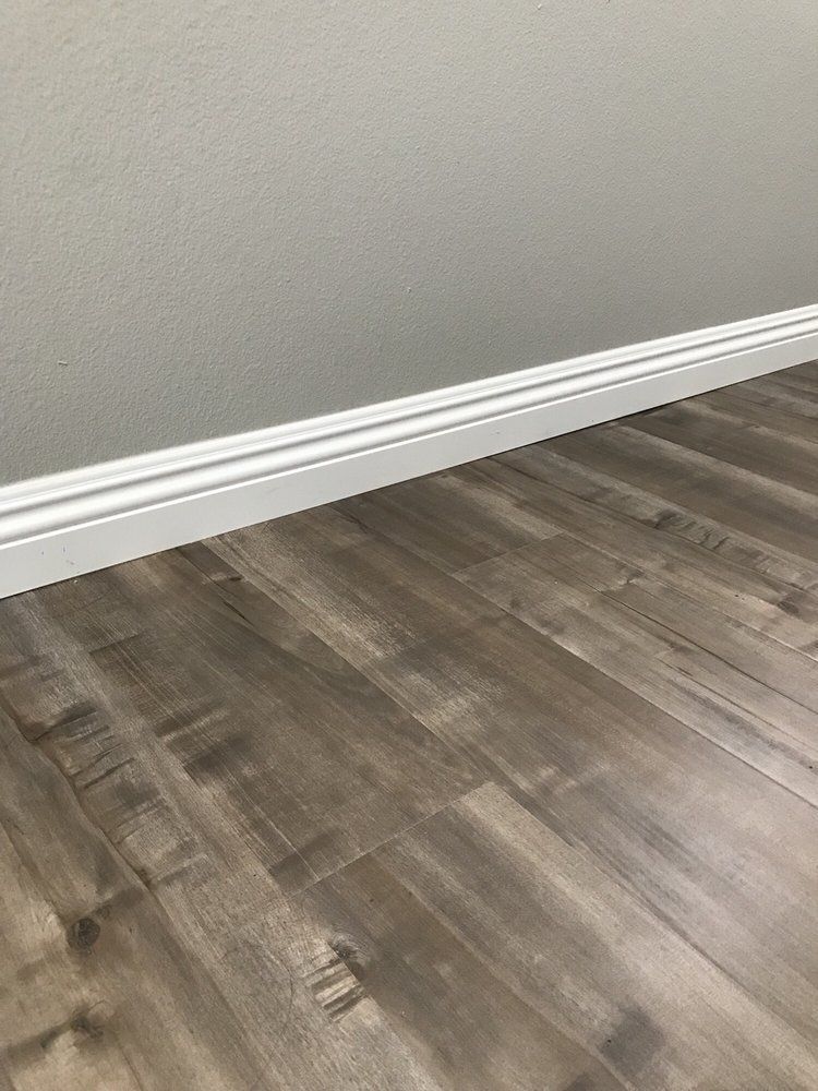 A view of grey wood-look laminate flooring meeting a white decorative baseboard against a light grey wall.