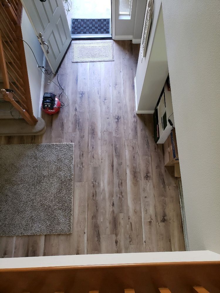 A high-angle view looking down at a wooden hallway floor with a gray area rug, leading to an open doorway.