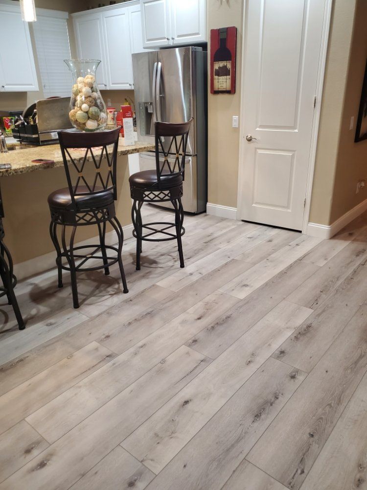 A modern kitchen featuring light wood-look flooring, a granite breakfast bar with two dark stools, and stainless fridge.