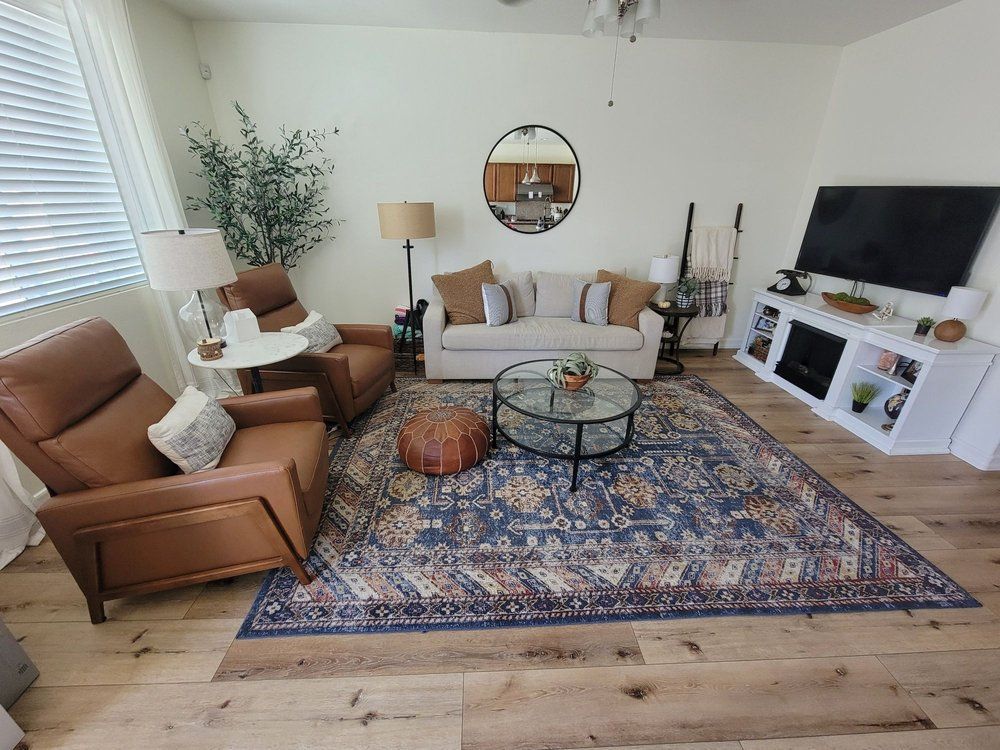A living room with a beige sofa, two brown leather armchairs, a blue patterned area rug, a glass coffee table, and a TV.