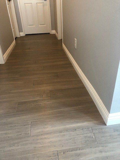 A hallway with grey walls, white baseboards, and light-colored, wood-grain plank flooring leading to a closed white door.