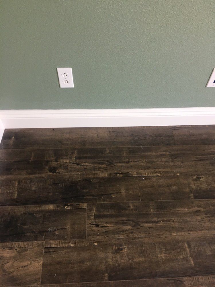 A white electrical outlet on a sage green wall above dark brown wood-look flooring and a white baseboard.