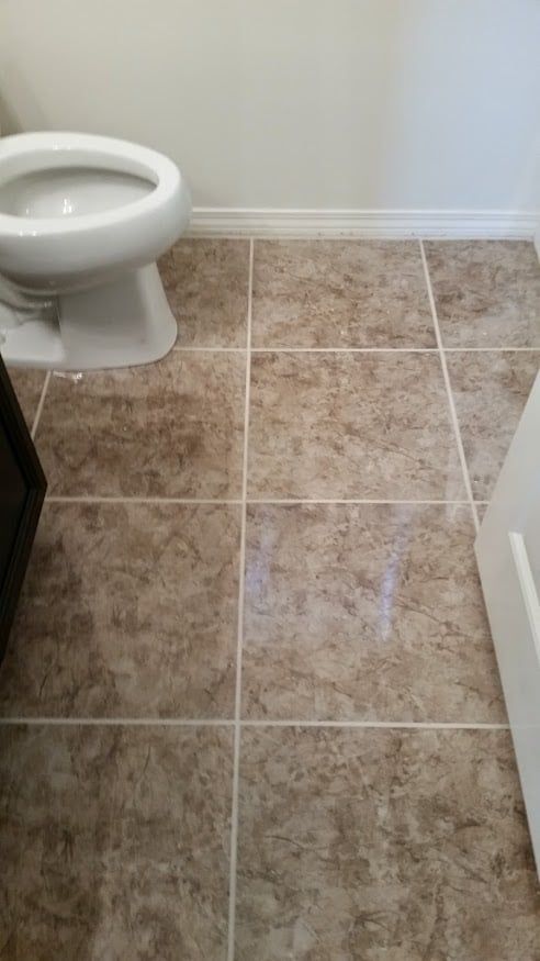 A bathroom floor featuring light brown, mottled square tiles with white grout, next to a white toilet base.