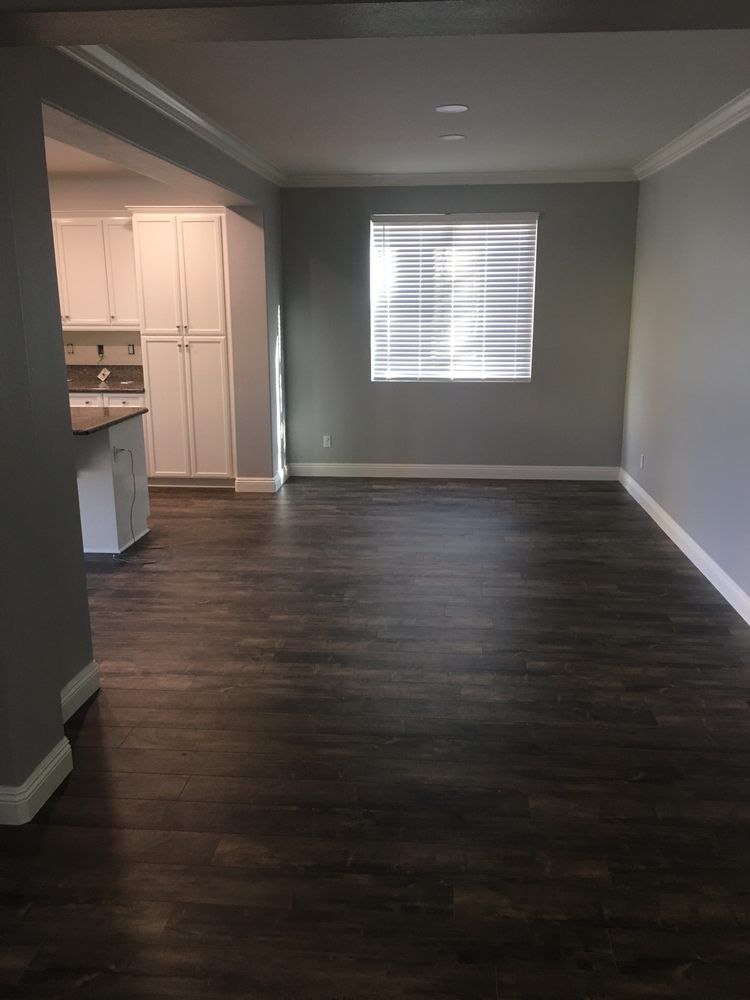 A room with dark wood floors, gray walls, white trim, and a window with blinds, opening into a white-cabinet kitchen.
