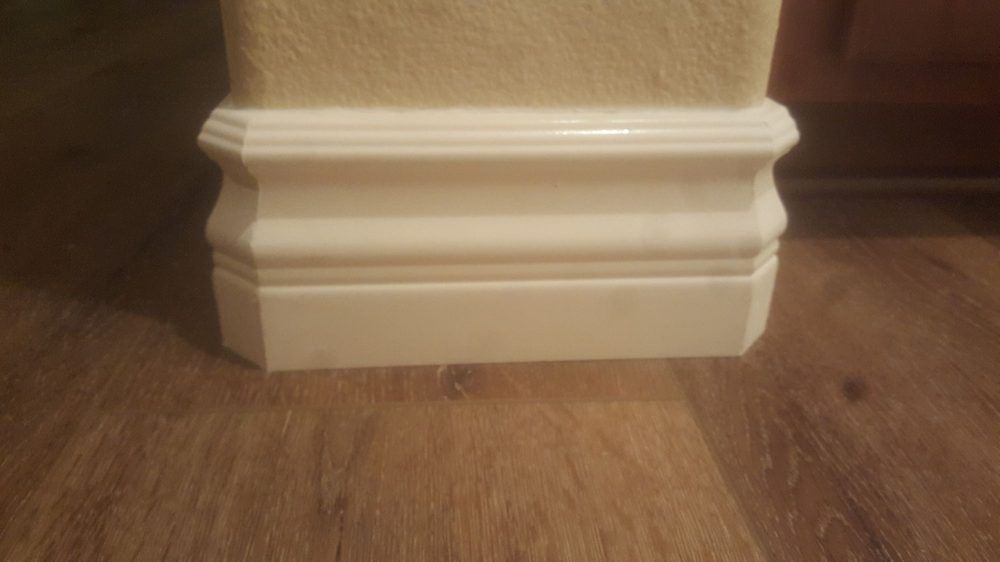 A close-up of decorative white baseboard molding installed on a textured wall against brown wood-look flooring.