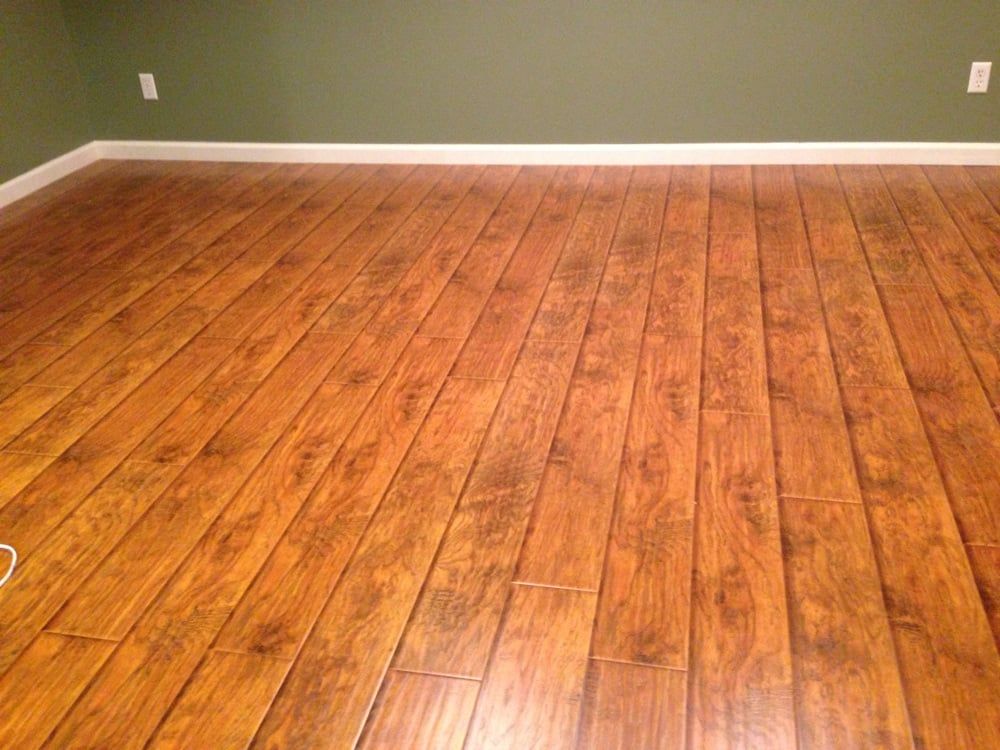 Polished, medium-toned wood plank flooring leading up to a plain sage green wall with white baseboards and wall outlets.