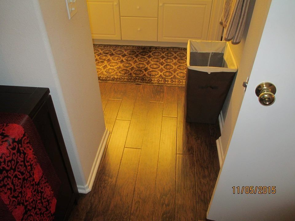 A doorway looks into a bathroom with wood-look flooring, a patterned rug, white vanity cabinets, and a tall laundry hamper.