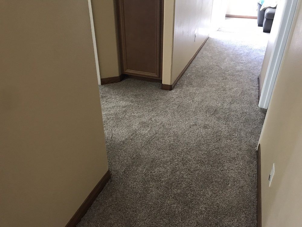 A hallway with beige walls, brown trim, and wall-to-wall grey carpet leading to an open doorway at the far end.