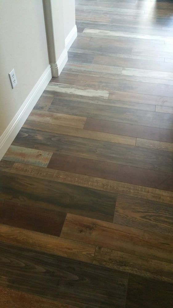 A close-up view of rustic-style wood-look flooring planks in varying shades of brown, gray, and tan next to a white wall.