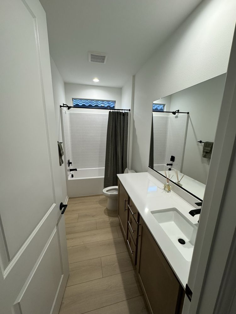 A bathroom with a long vanity featuring a white countertop, a mirror, wood cabinets, a toilet, and a tiled tub area.