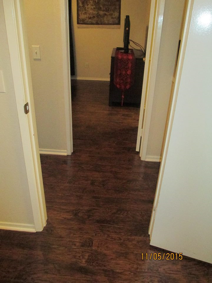 A view through a doorway into a hallway with dark wood-look laminate flooring, leading to a room with a decorative table.
