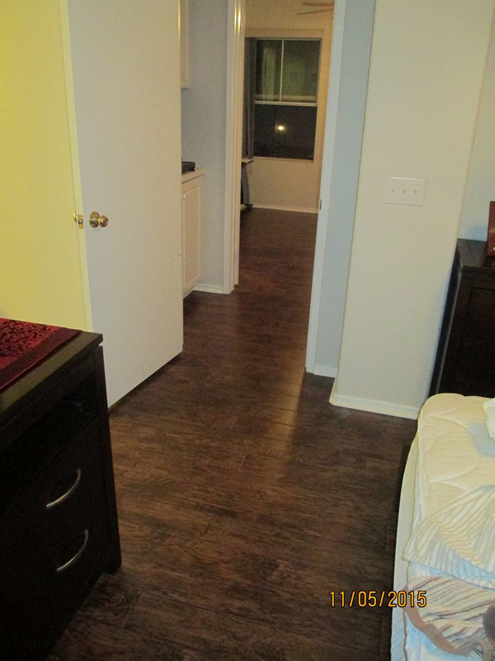 A hallway with dark wood-look flooring leads to a room with a window, flanked by a yellow wall and white door.