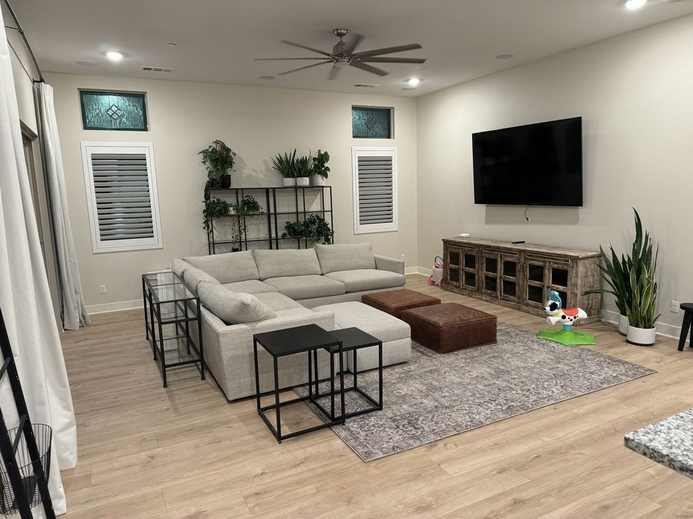 A living room with a large light gray sectional, two black side tables, a brown ottoman, and a TV mounted on a wall.