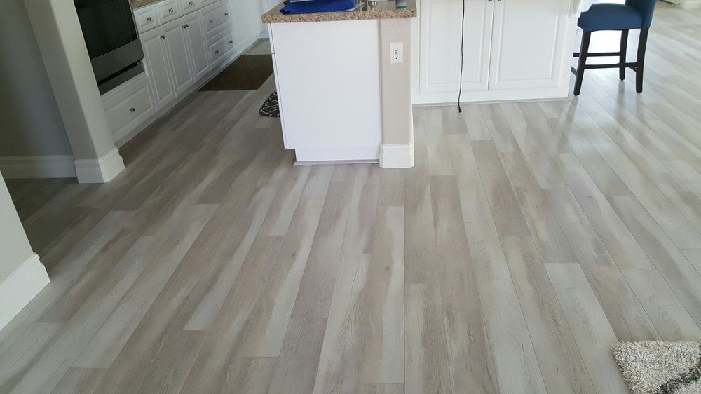 Light wood-look flooring stretches across a kitchen area with white cabinets, an island, and a single blue bar stool.