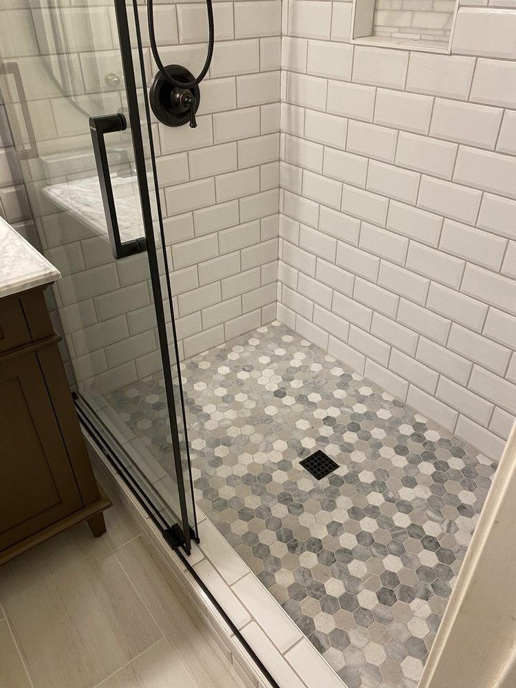 A walk-in shower with white subway tile walls, a gray and white hexagonal tile floor, and a black-framed glass door.