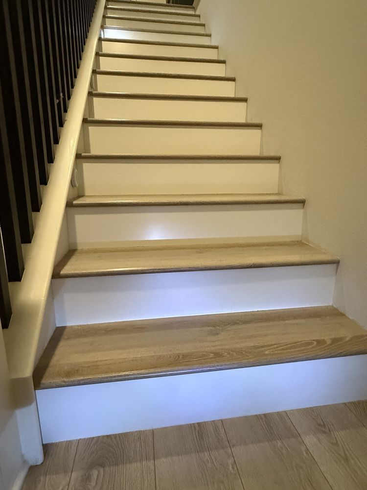 A set of stairs with light wood-grain treads and white risers, viewed from the bottom looking up toward a dark railing.