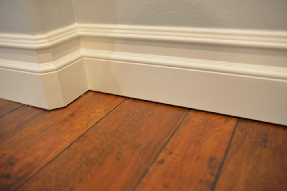 A close-up of white wooden baseboard trim meeting at a corner above dark brown wooden flooring.