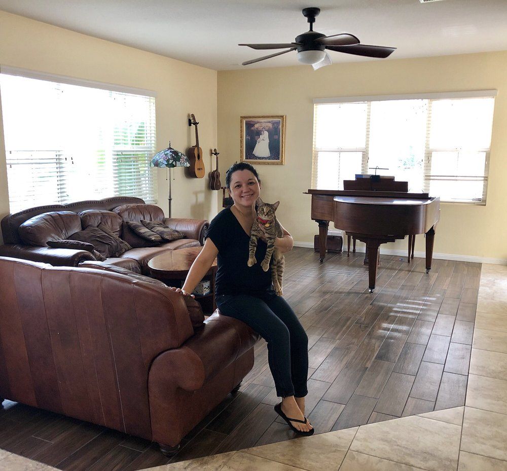 A person holding a cat while sitting on a brown leather sofa in a living room with a piano, guitars, and wood flooring.