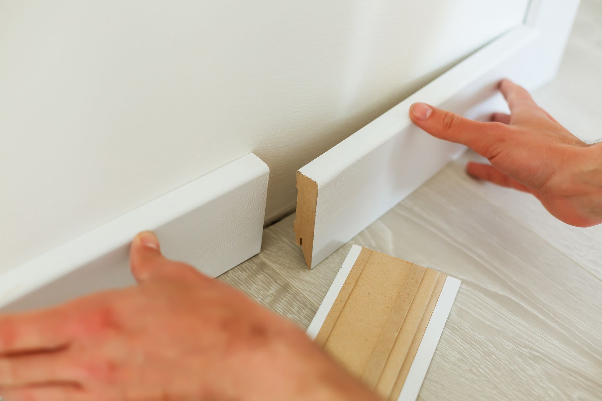 Hands installing white baseboard trim along a wall, with a piece lying on the floor.