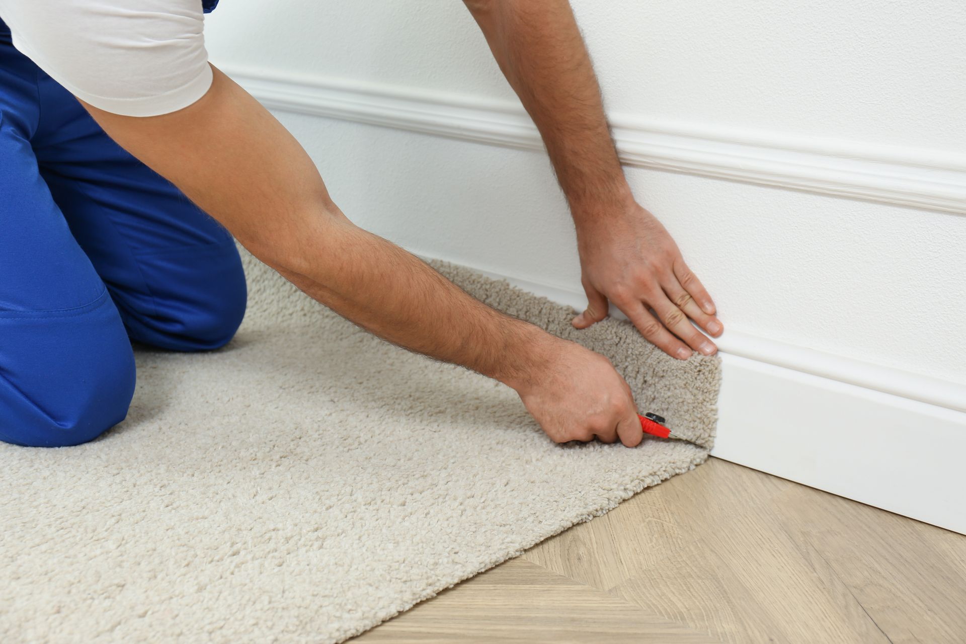Person kneeling, cutting carpet against a white wall and baseboard.