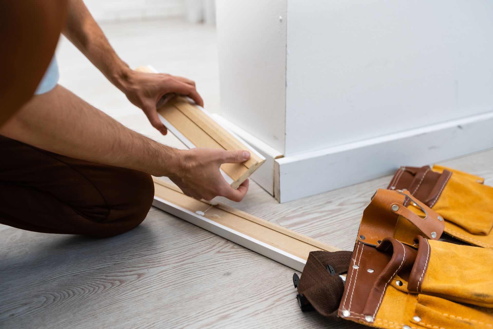 Person installing wooden trim near a white wall and tool belt on floor.