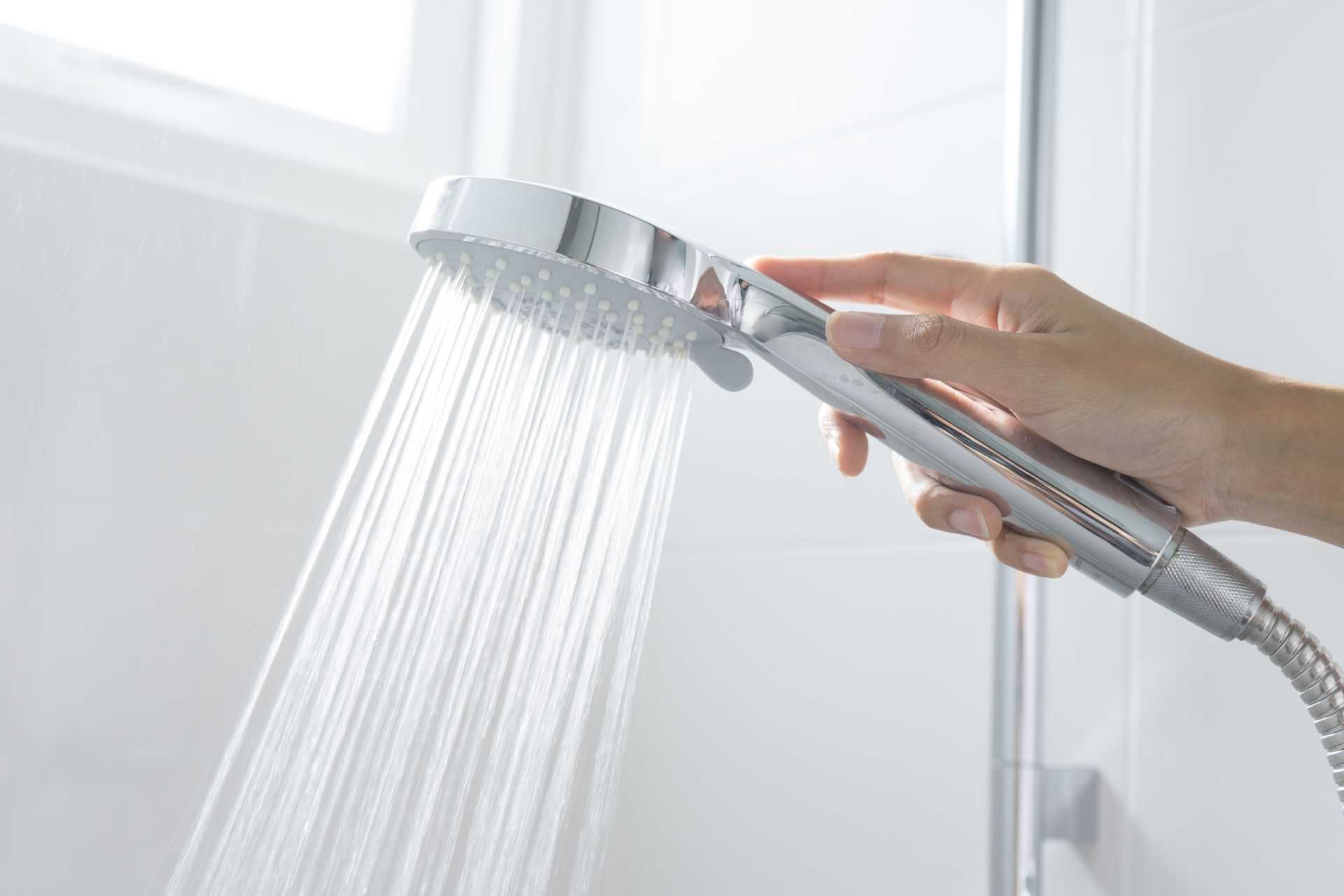 Hand holding a chrome shower head with water streaming down in a white tiled bathroom.