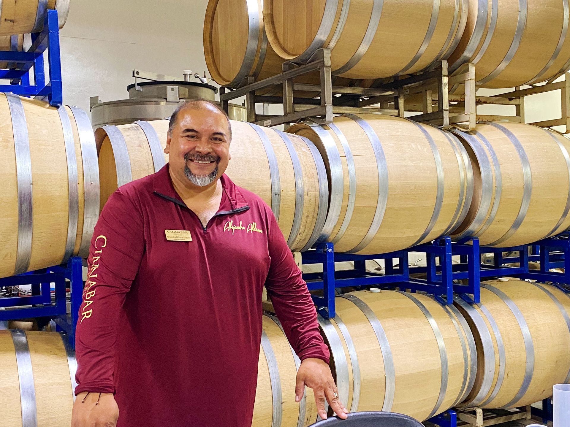 A smiling person in a maroon long-sleeved shirt standing in a cellar filled with stacks of wooden wine barrels.