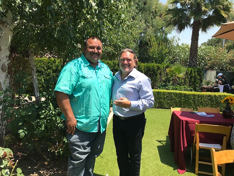 Two men standing side-by-side in a sunlit garden, one in a turquoise shirt and one in a light blue dress shirt.